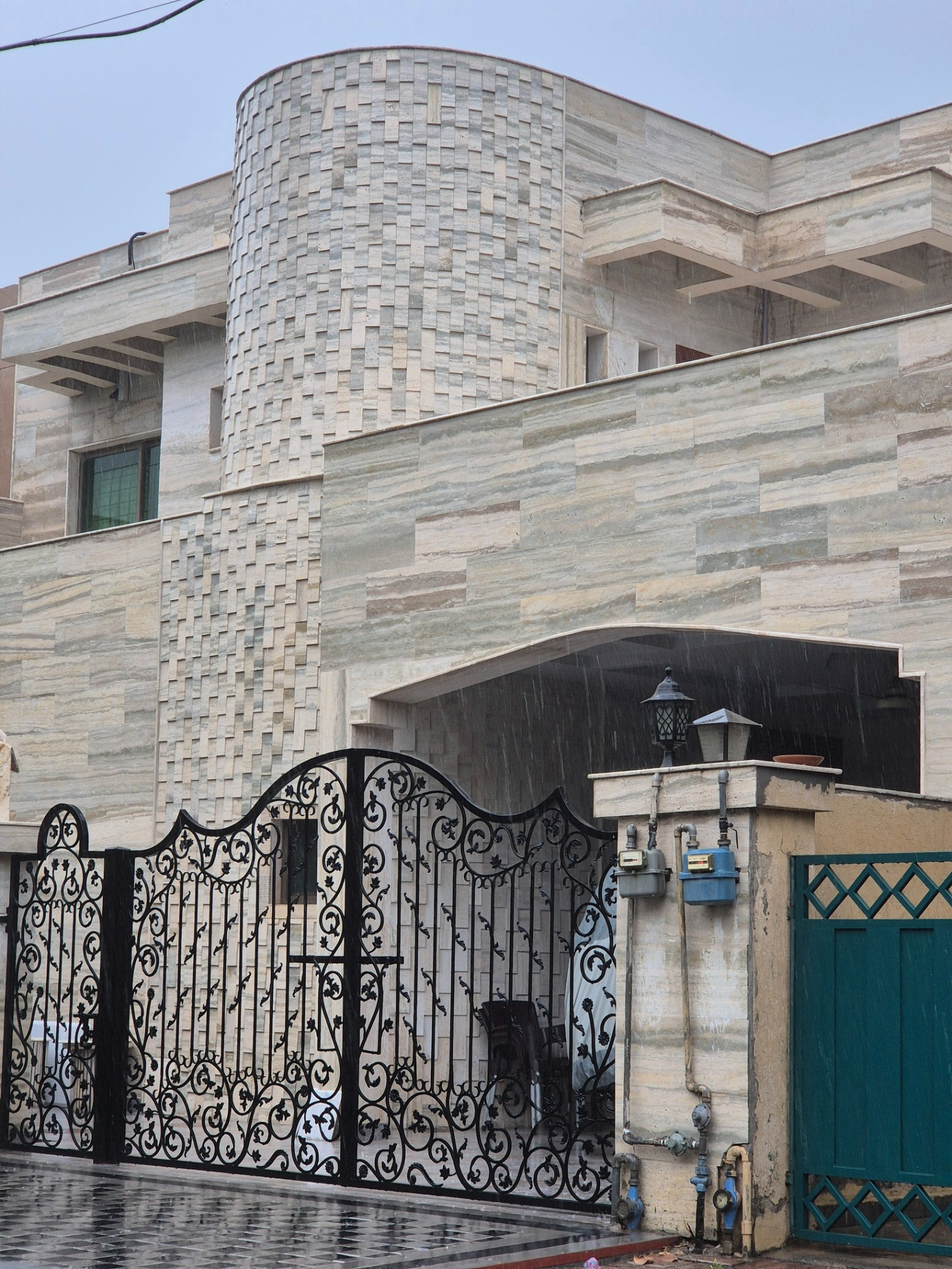 Modern house facade clad in travertine with a curved tower and ornate iron gate