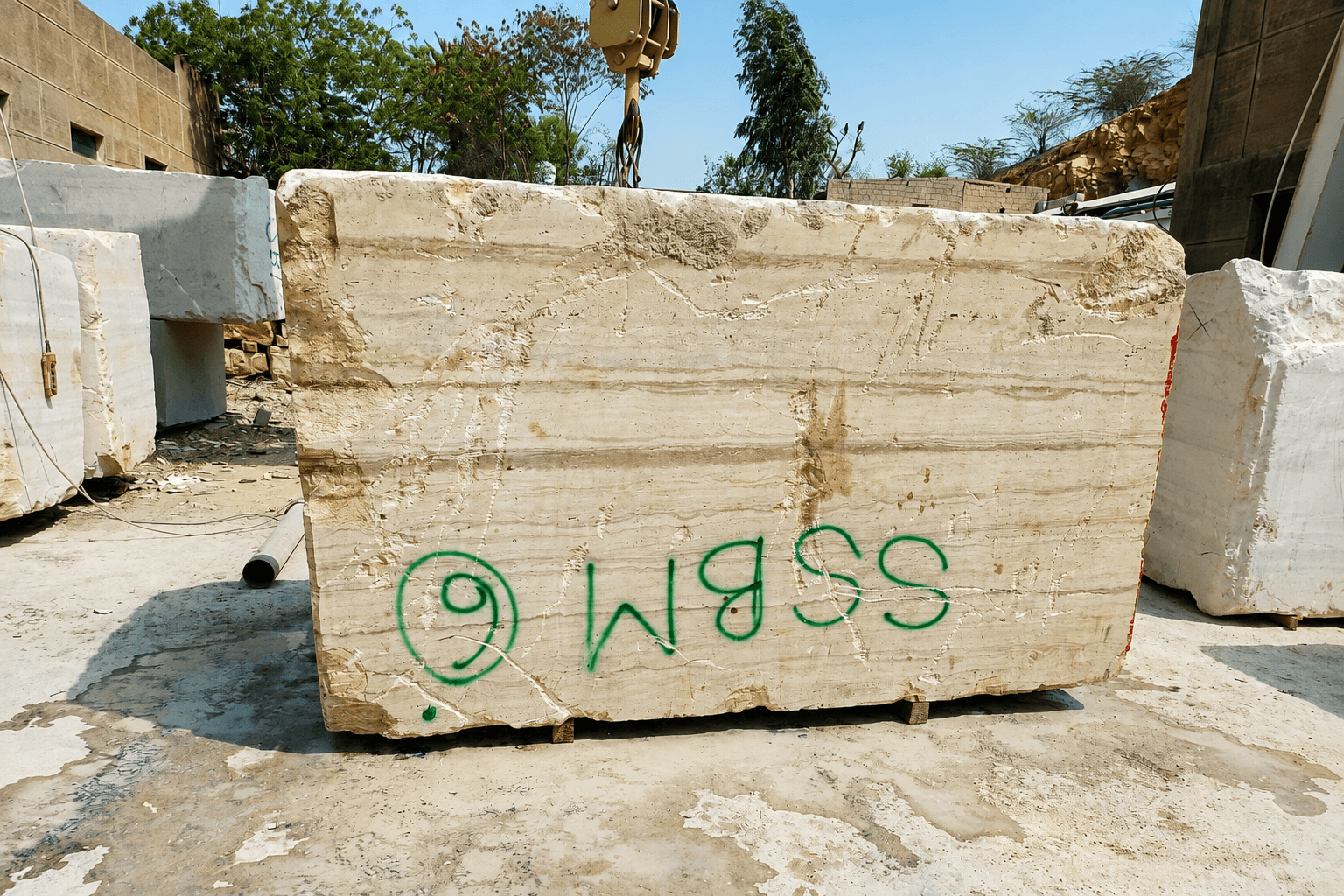 Raw travertine block with quarry identification markings and a crane hook suspended above, in the quarry yard